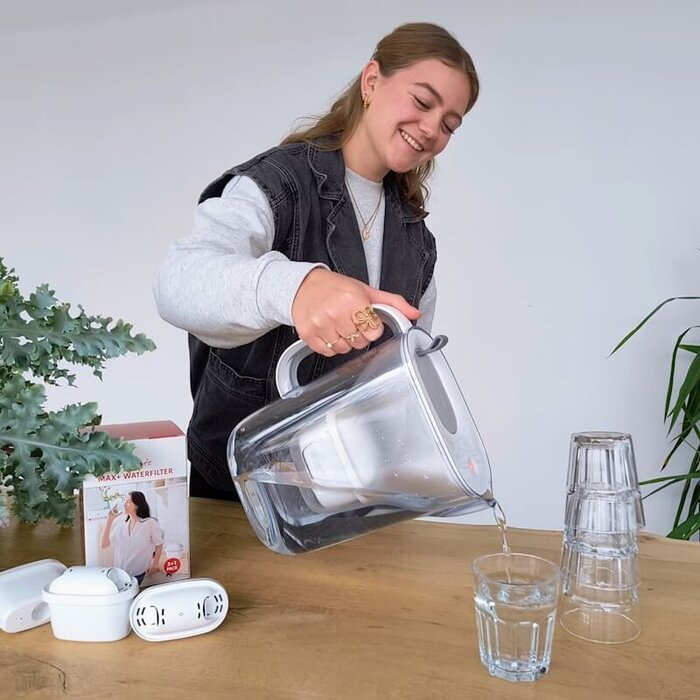 Een lachende vrouw giet water uit een filterkan in een glas op een houten tafel, met filterdozen in de buurt.