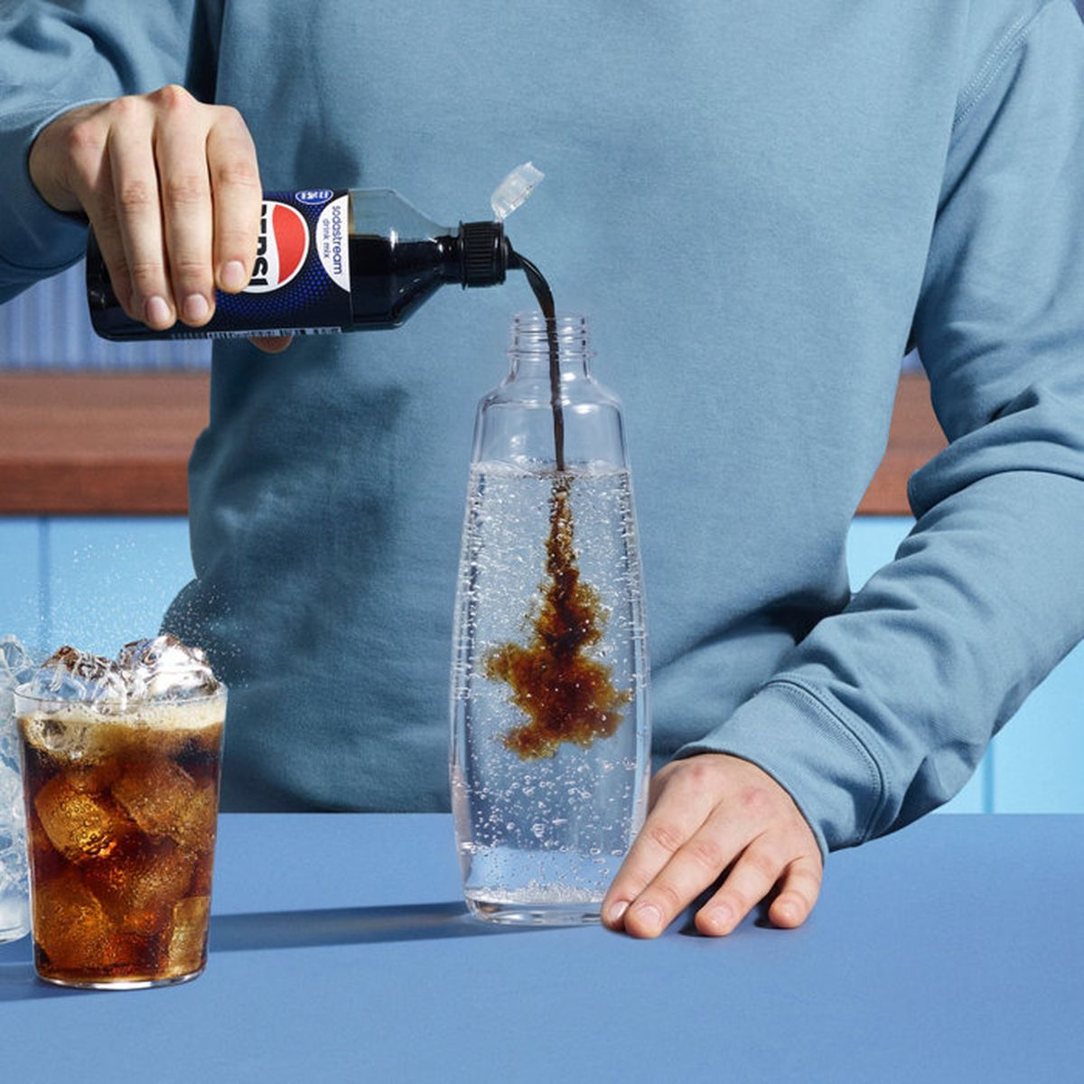 Persona vierte jarabe en una botella de agua con gas, con un vaso de refresco y hielo.