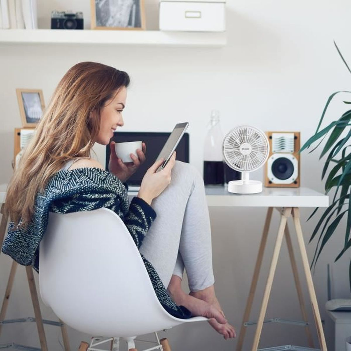Donna seduta su sedia alla scrivania, tiene tablet e tazza, con un piccolo ventilatore bianco accanto.