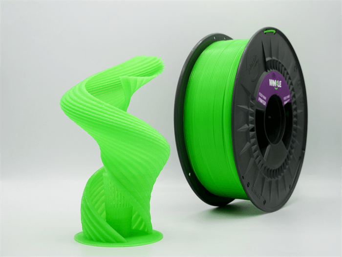 Una escultura espiral verde brillante impresa en 3D junto a un carrete de filamento verde en una superficie blanca.