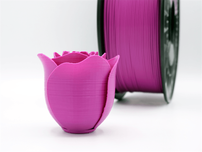 Una tazza a tulipano rosa stampata in 3D si trova di fronte a una bobina di filamento coordinata su sfondo bianco.