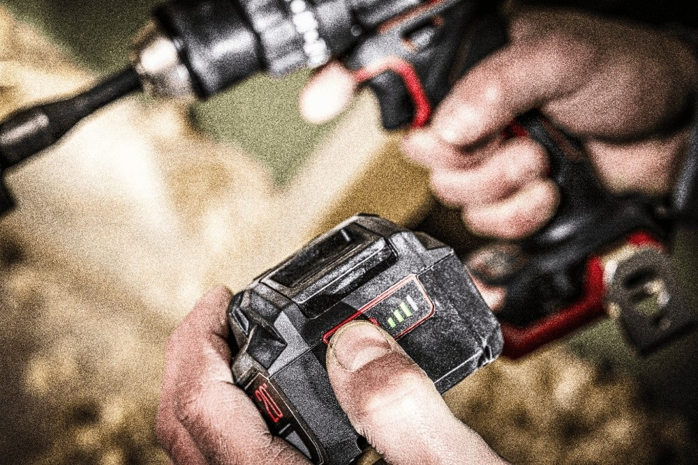 Persona che tiene una batteria per trapano con indicatore di carica verde, con il trapano sullo sfondo.