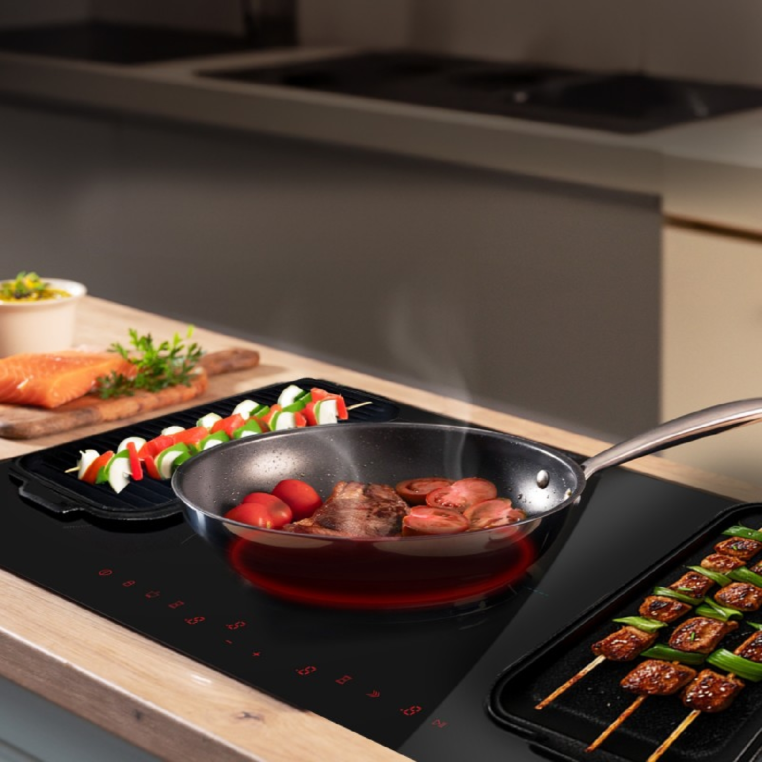 Carne e pomodori friggono in padella su piano induzione, con spiedini e salmone accanto.