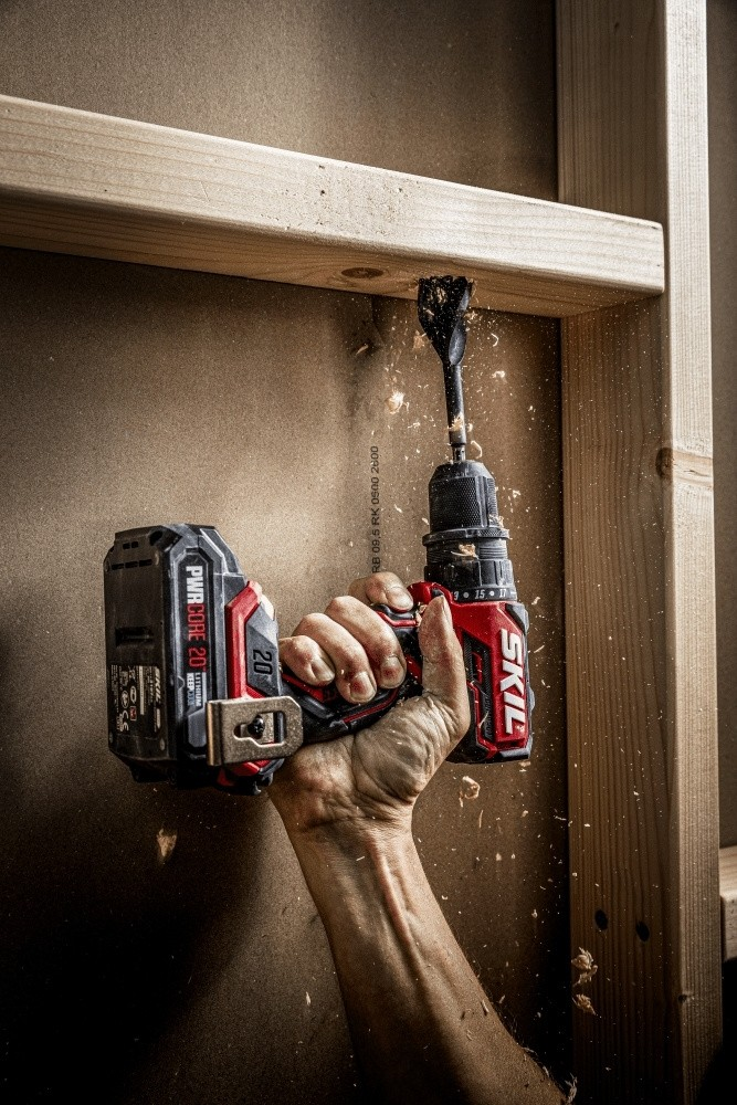 Una mano usa un trapano a batteria Skil rosso per fare un foro in una trave di legno con trucioli.