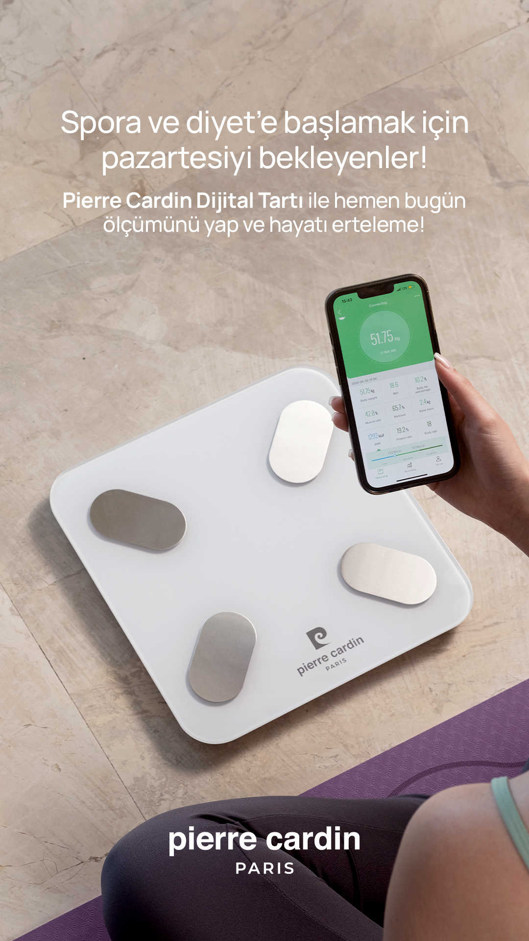 Bir kişi karolu zeminde beyaz dijital tartı yanında kilo verilerini gösteren telefon tutuyor.
