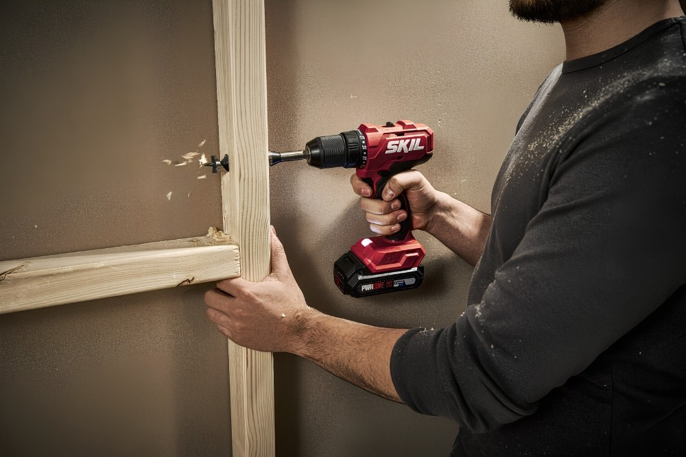 Un uomo fora una trave di legno usando un trapano a batteria rosso.
