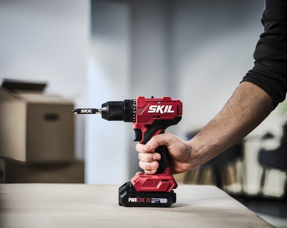 Una persona tiene in mano un trapano a batteria Skil rosso e nero su un tavolo di legno, con scatoloni sfocati.