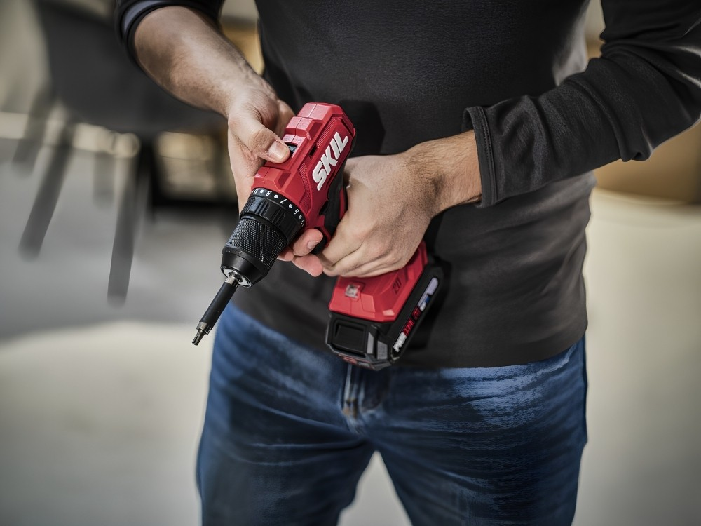Una persona tiene in mano un trapano a batteria Skil rosso e nero, con maglia scura e jeans al chiuso.
