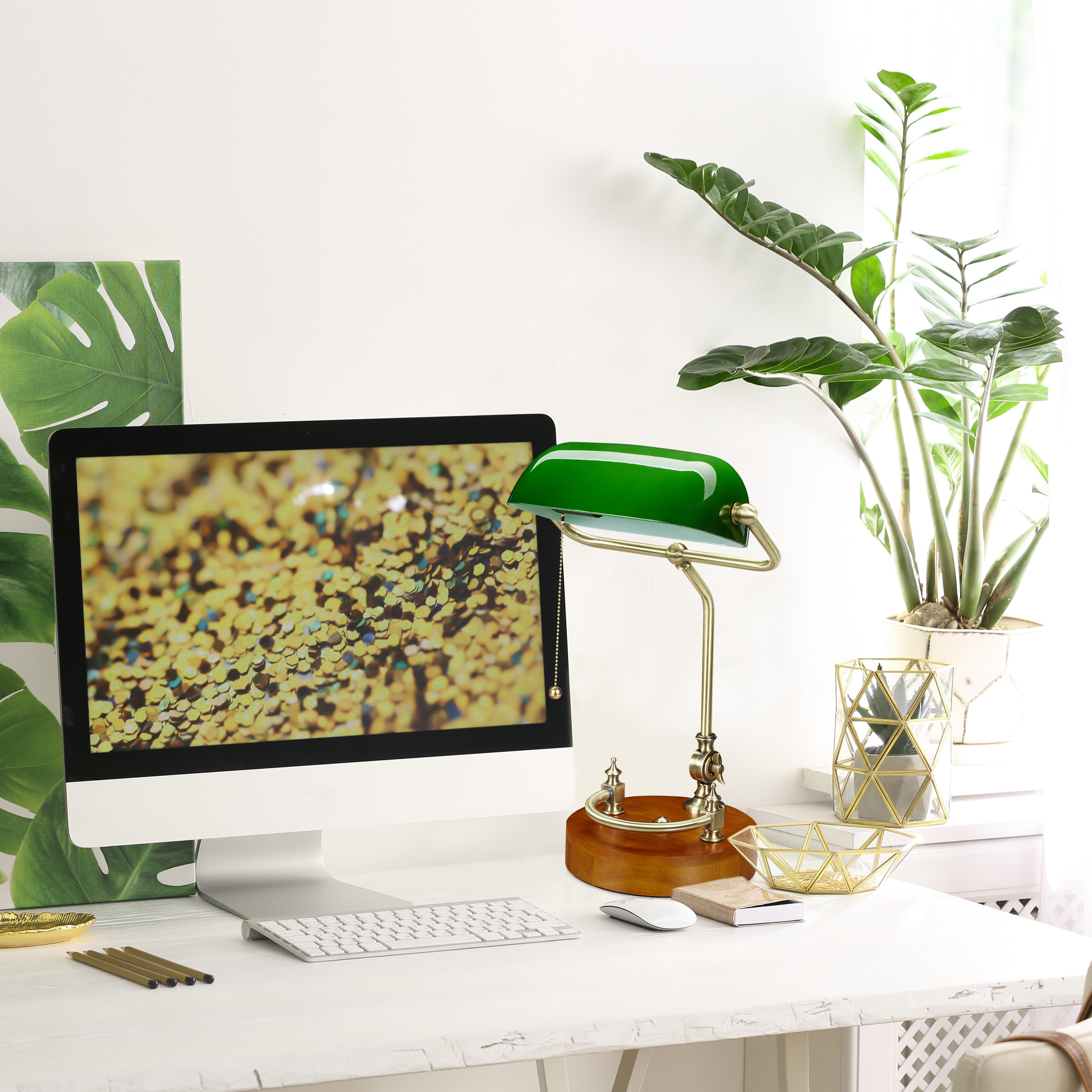 Ein weißer Schreibtisch mit einem Monitor, einer grünen Bankerlampe, einer Tastatur und Pflanzen im Hintergrund.