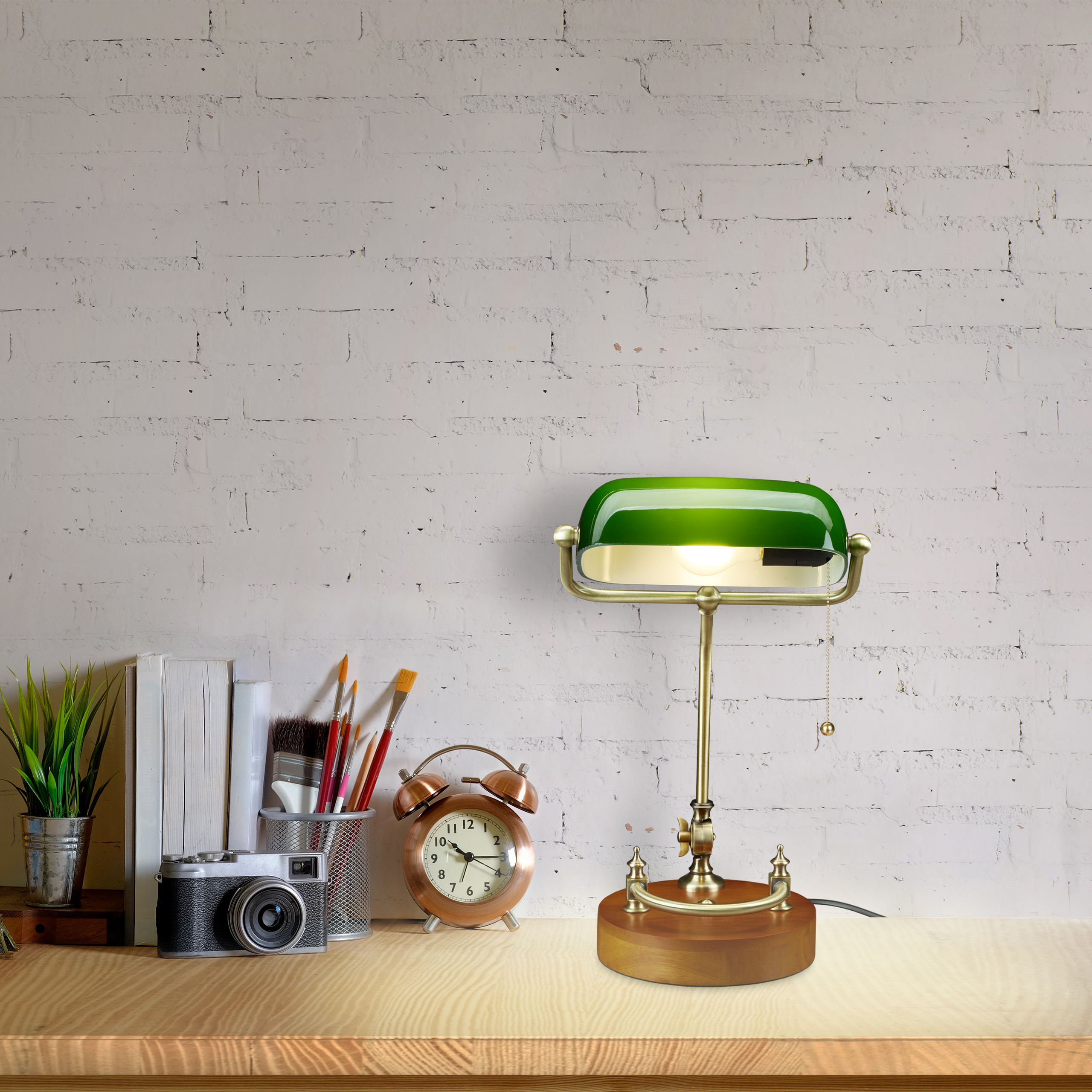 Eine grüne Bankerlampe steht auf einem Holztisch, neben Büchern, einer Kamera, einer Uhr und einer Pflanze vor einer Backsteinwand.