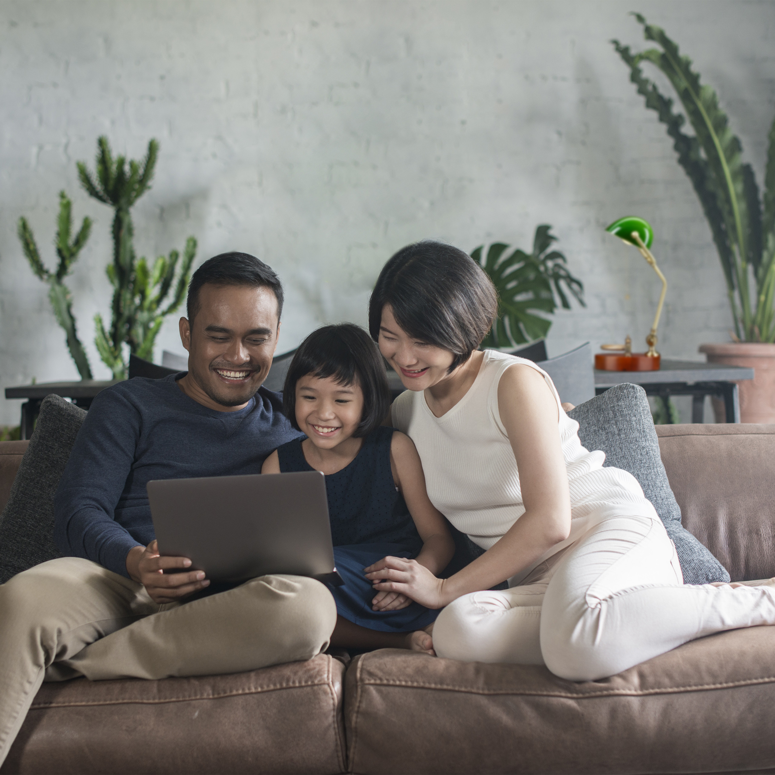 Eine glückliche dreiköpfige Familie sitzt auf einem braunen Sofa und lächelt auf einen Laptop.