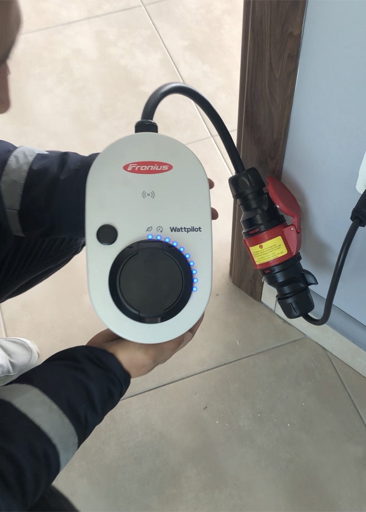 Bir kişi, takılı siyah bir kabloya sahip beyaz bir Fronius Wattpilot EV şarj cihazı tutuyor.