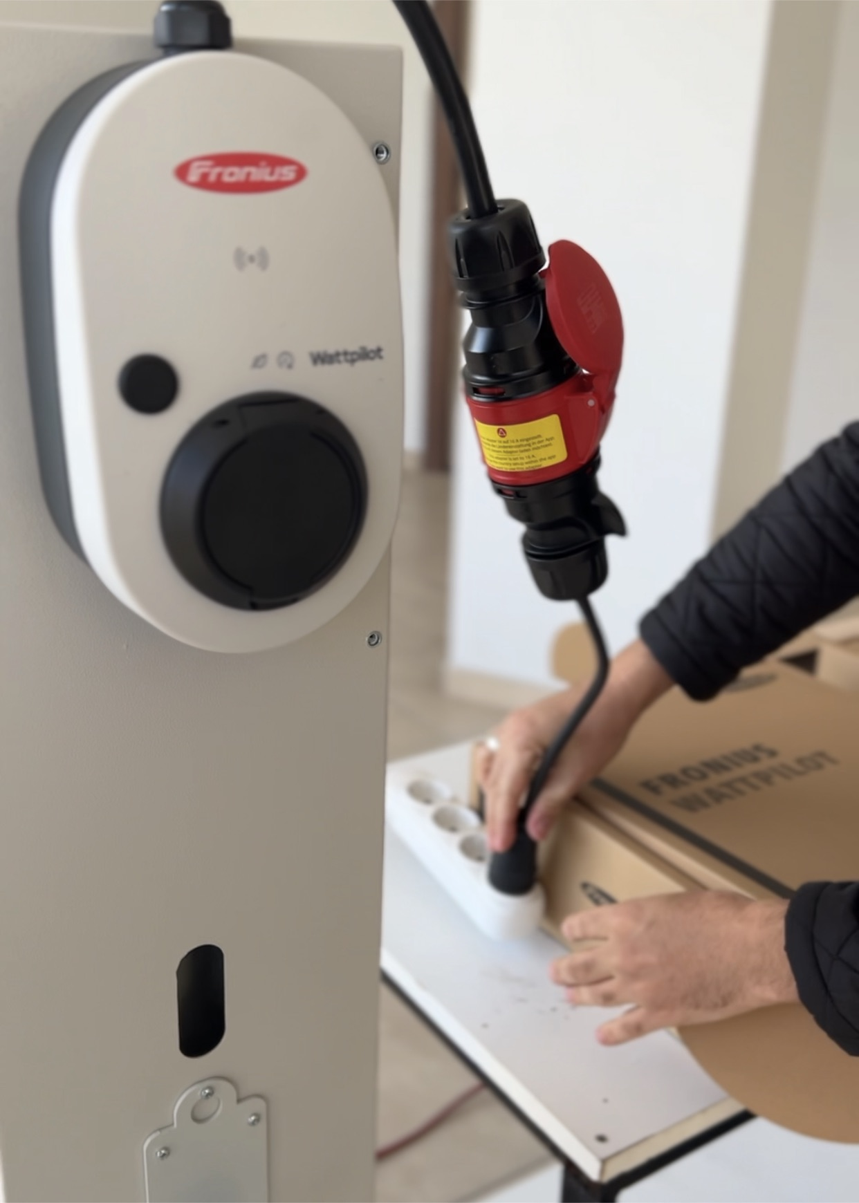 Bir kişi, Fronius Wattpilot ünitesinin yanında siyah bir şarj kablosunu beyaz bir uzatma kablosuna takıyor.