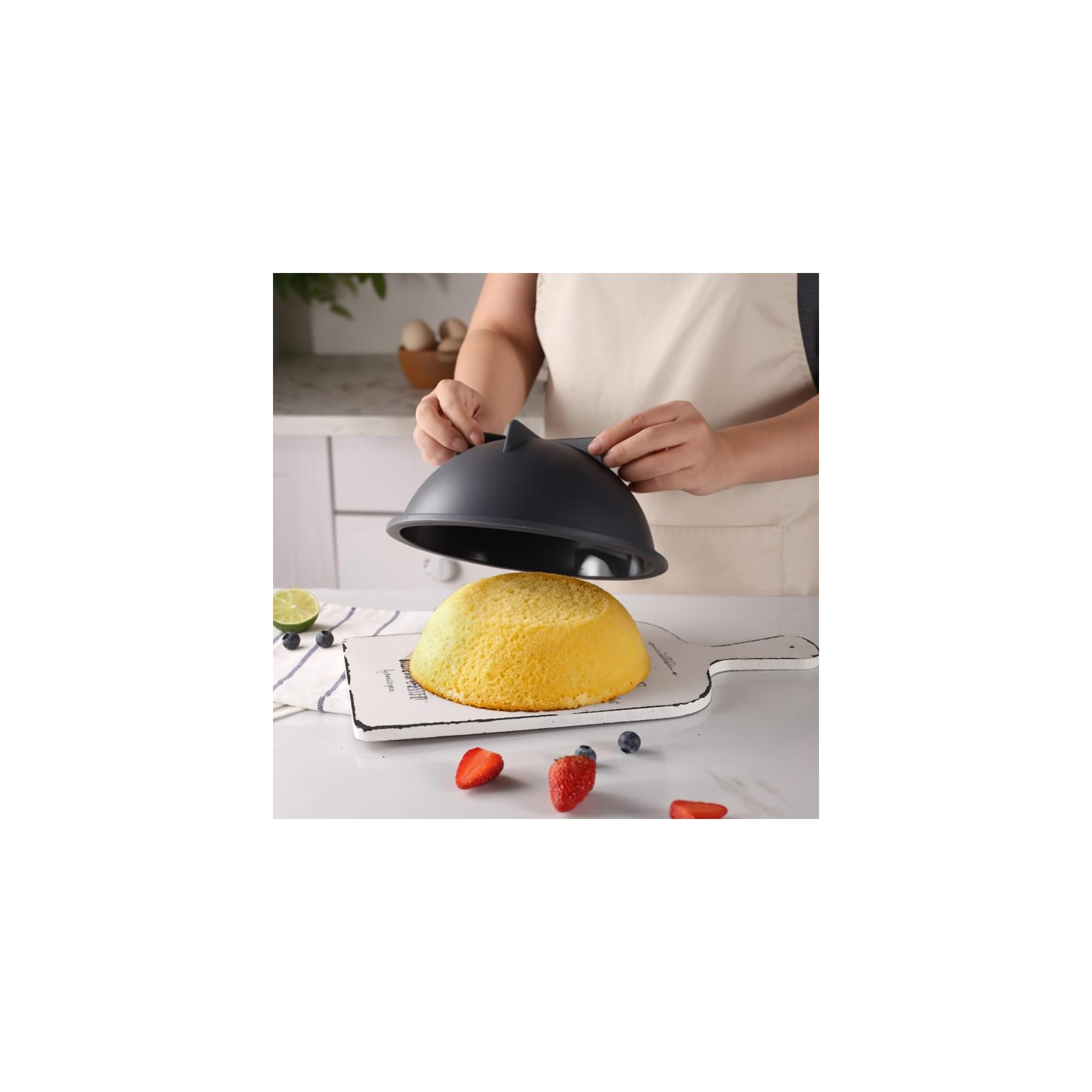 Eine Person mit Schürze hebt eine dunkle Kuppelhaube von einem runden gelben Kuchen ab.