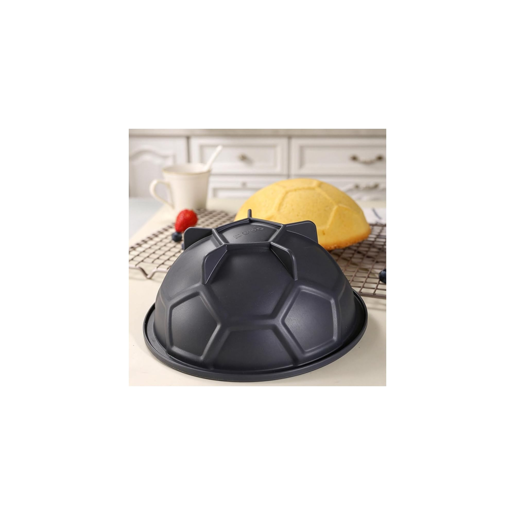Eine schwarze Fußball-Backform auf einem Tisch mit gebackenem Kuchen, Erdbeere und Tasse im verschwommenen Hintergrund.