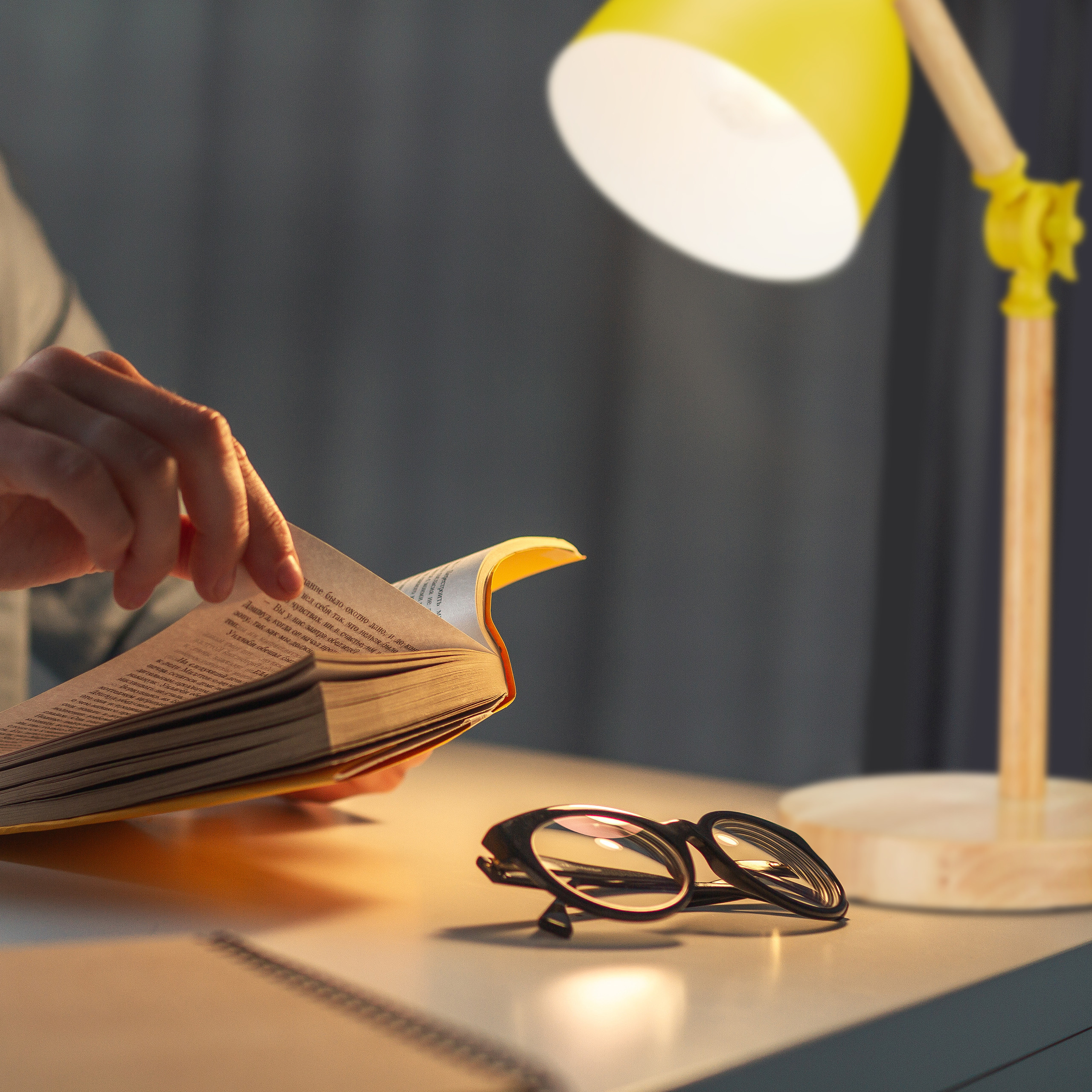 Person liest Buch am Tisch unter gelber Lampe neben Brille.