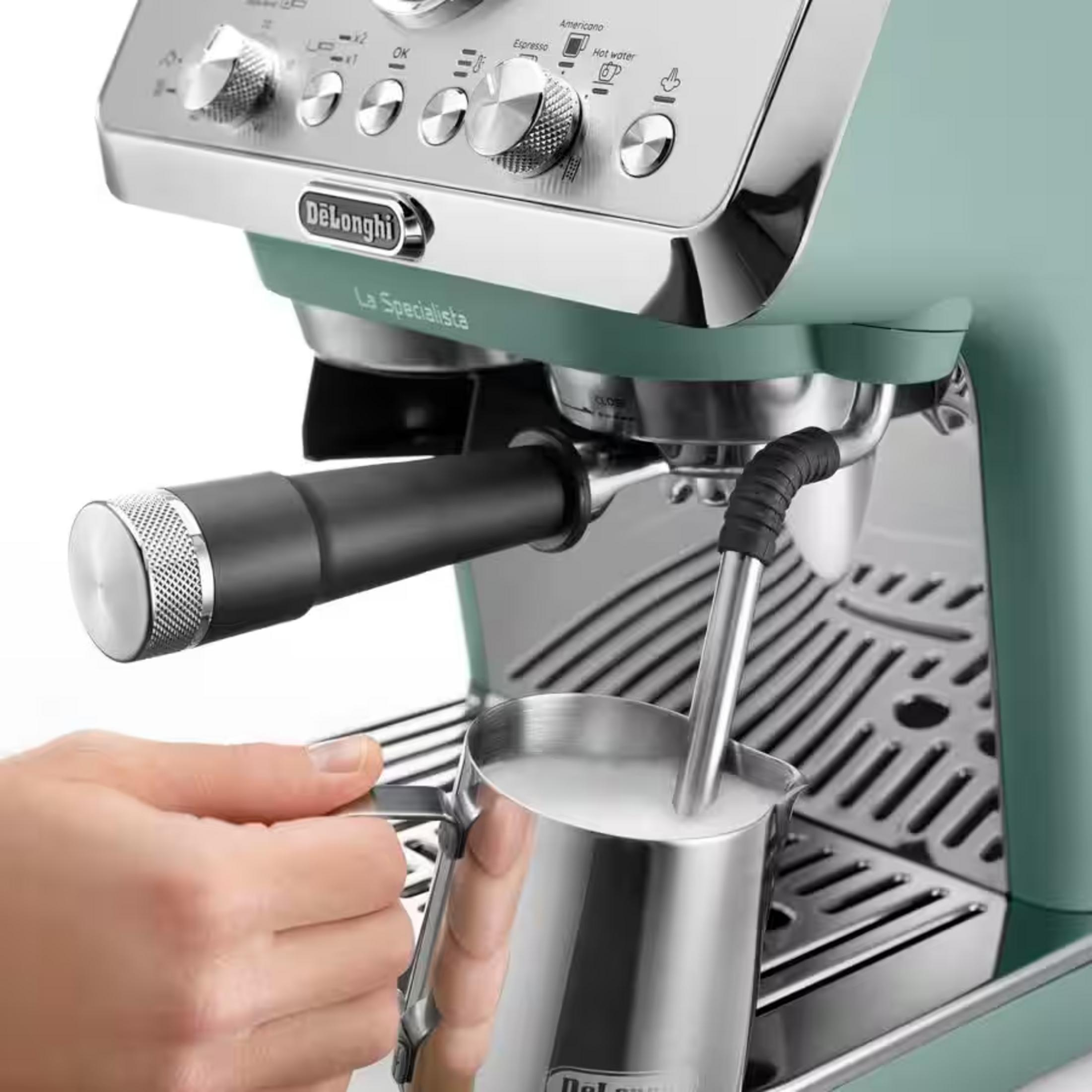 Yeşil bir DeLonghi kahve makinesinin buhar çubuğu altında süt sürahisi tutan bir el.