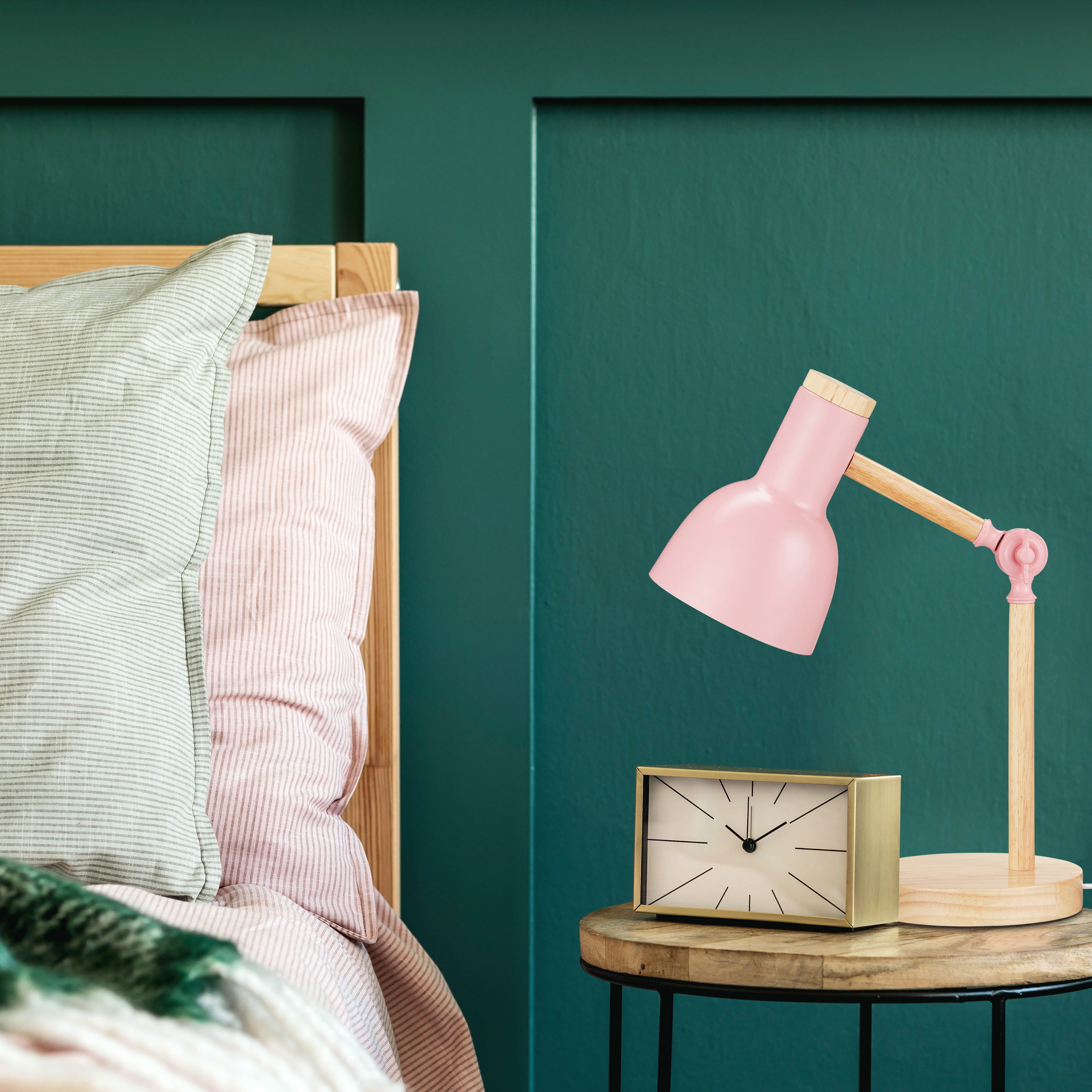 Eine rosa Schreibtischlampe und eine moderne Uhr stehen auf einem Nachttisch neben einem Bett vor einer grünen Wand.