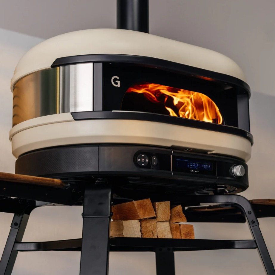 Ein cremefarbener und schwarzer Pizzaofen mit brennenden Flammen im Inneren steht auf einem schwarzen Metallständer mit gestapeltem Brennholz.