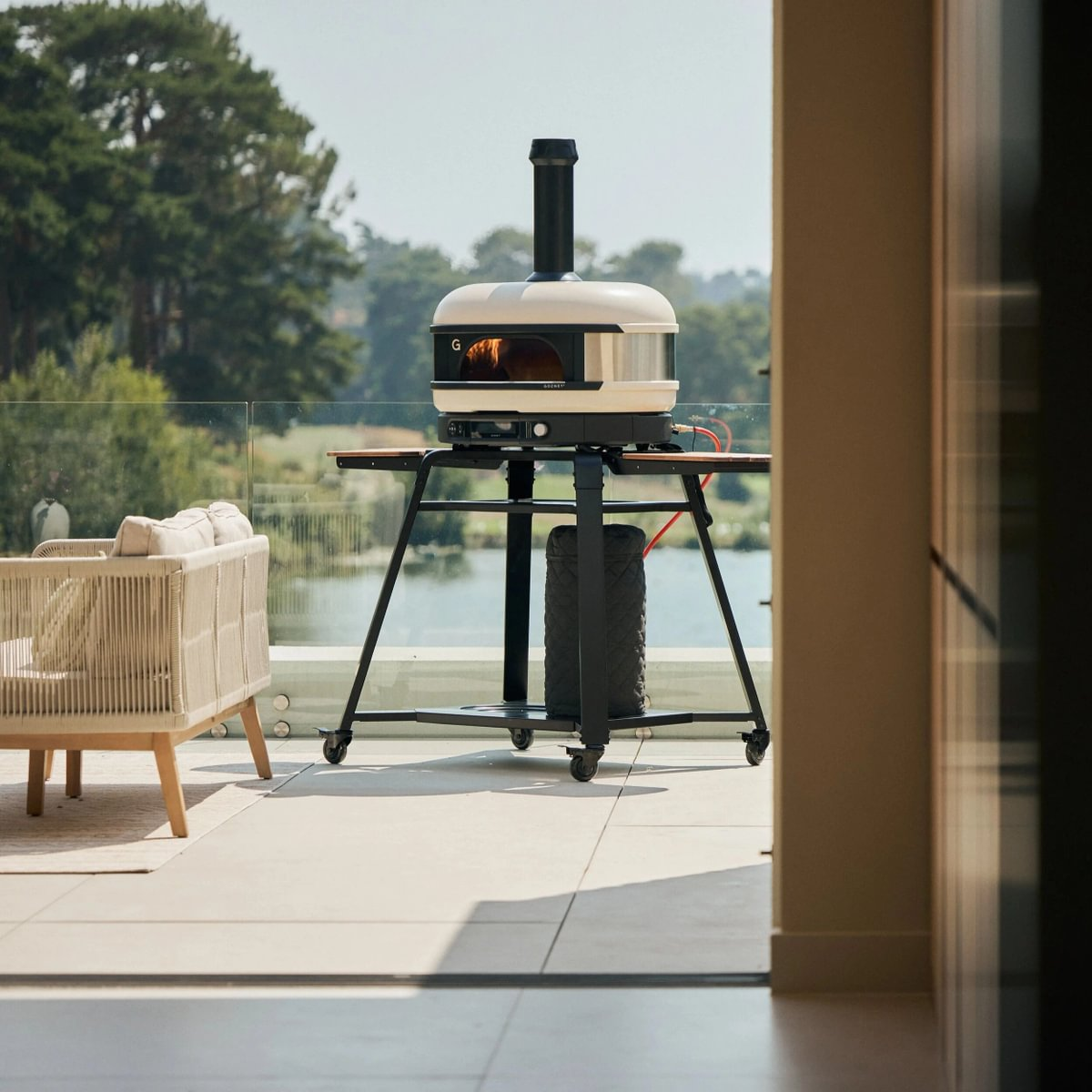Ein weißer Pizzaofen auf einem Metallwagen auf einem Balkon mit Blick auf See und Bäume.