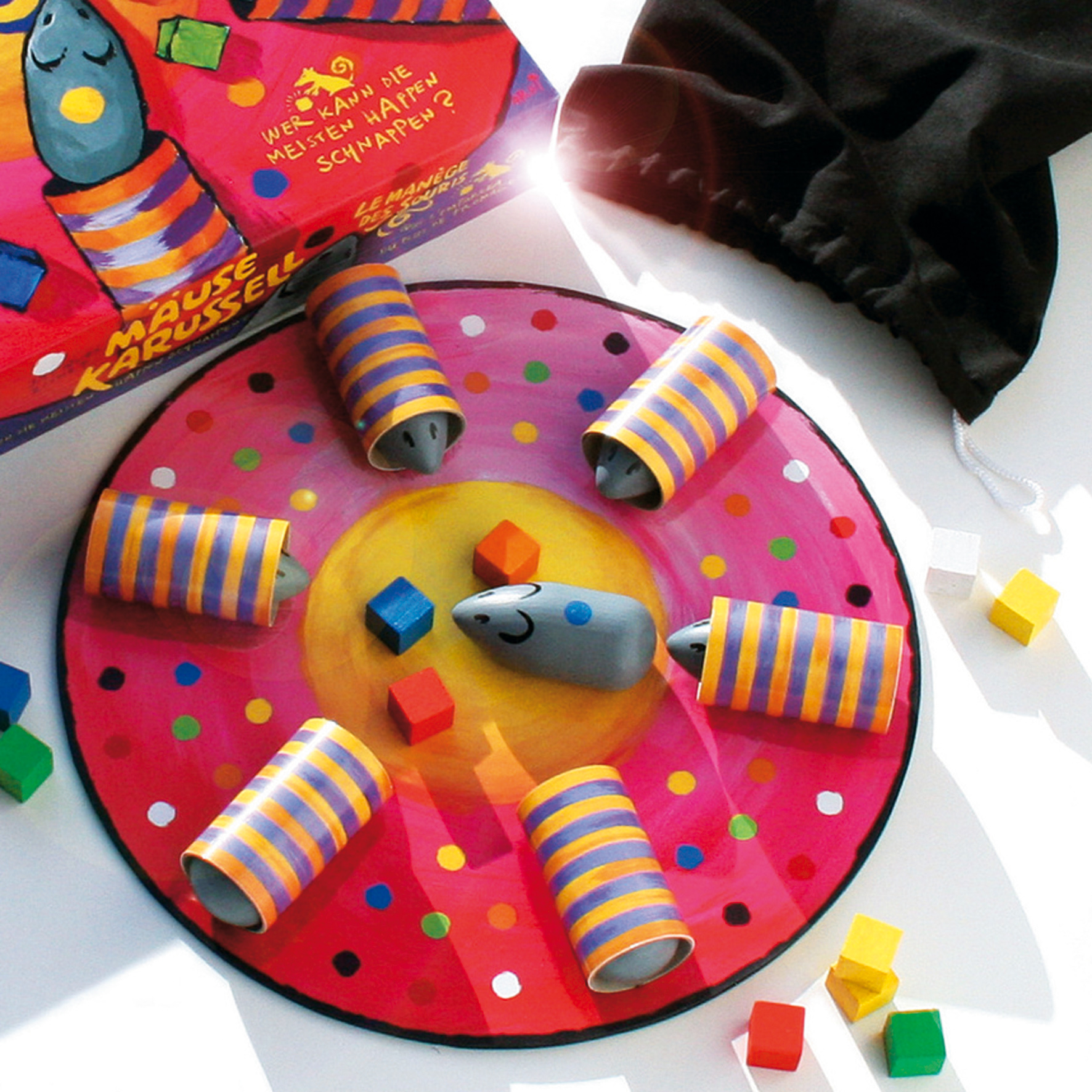 Ein buntes Brettspiel mit Mausfiguren in gestreiften Röcken, kleinen Holzwürfeln, Schachtel und schwarzem Stoffbeutel.