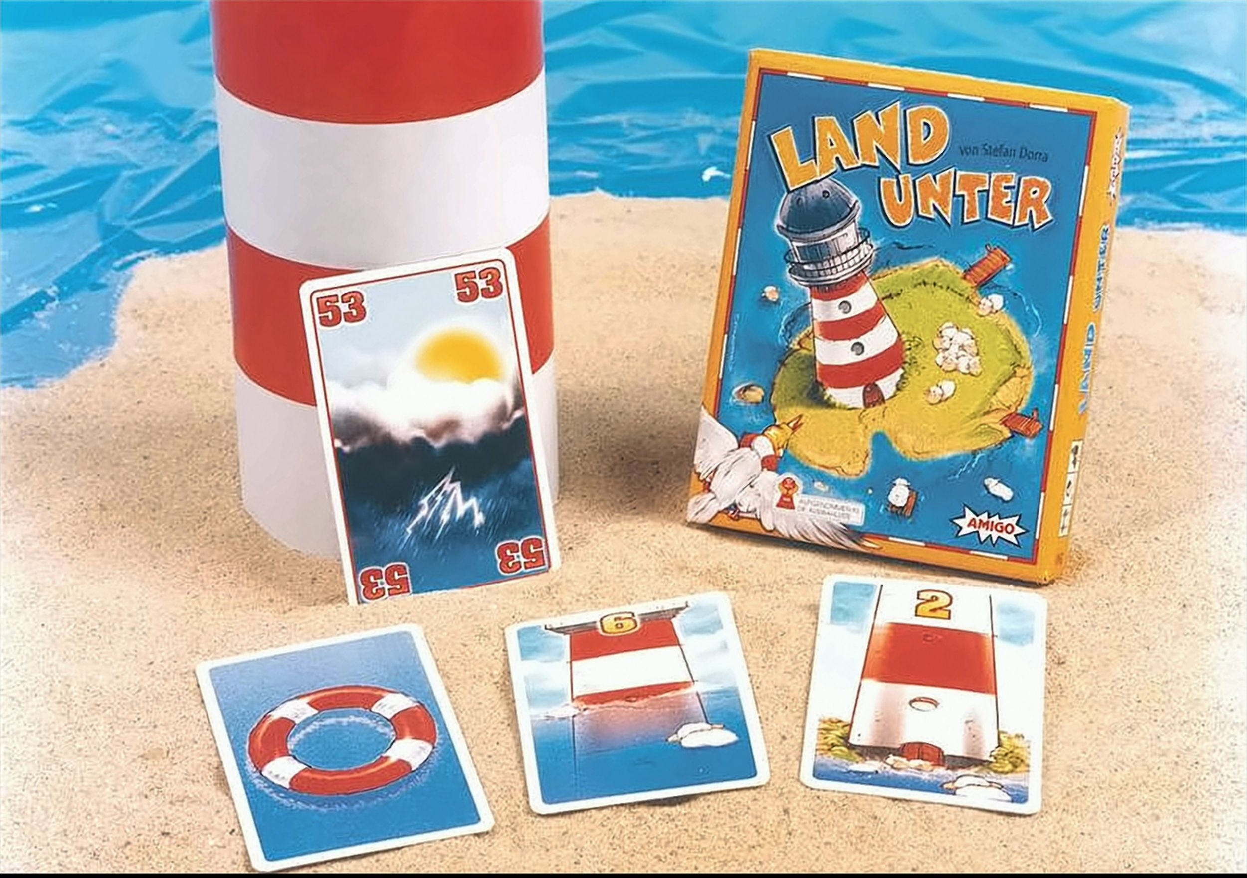 Das Brettspiel Land Unter mit Karten, Leuchtturm-Schachtel und einem Modell auf Sand am Wasser.