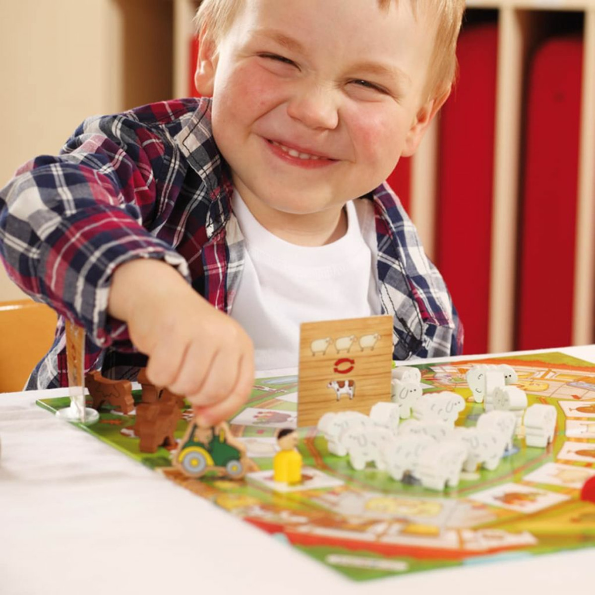 Ein lächelnder Junge im Karohemd spielt Brettspiel mit Spielzeugschafen und einem Traktor.