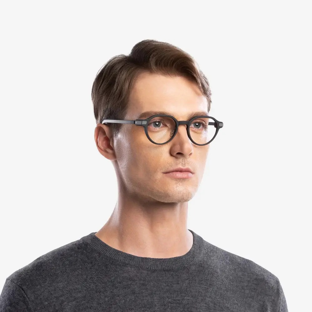 Un hombre con cabello castaño lleva gafas redondas negras y un suéter gris sobre fondo blanco.