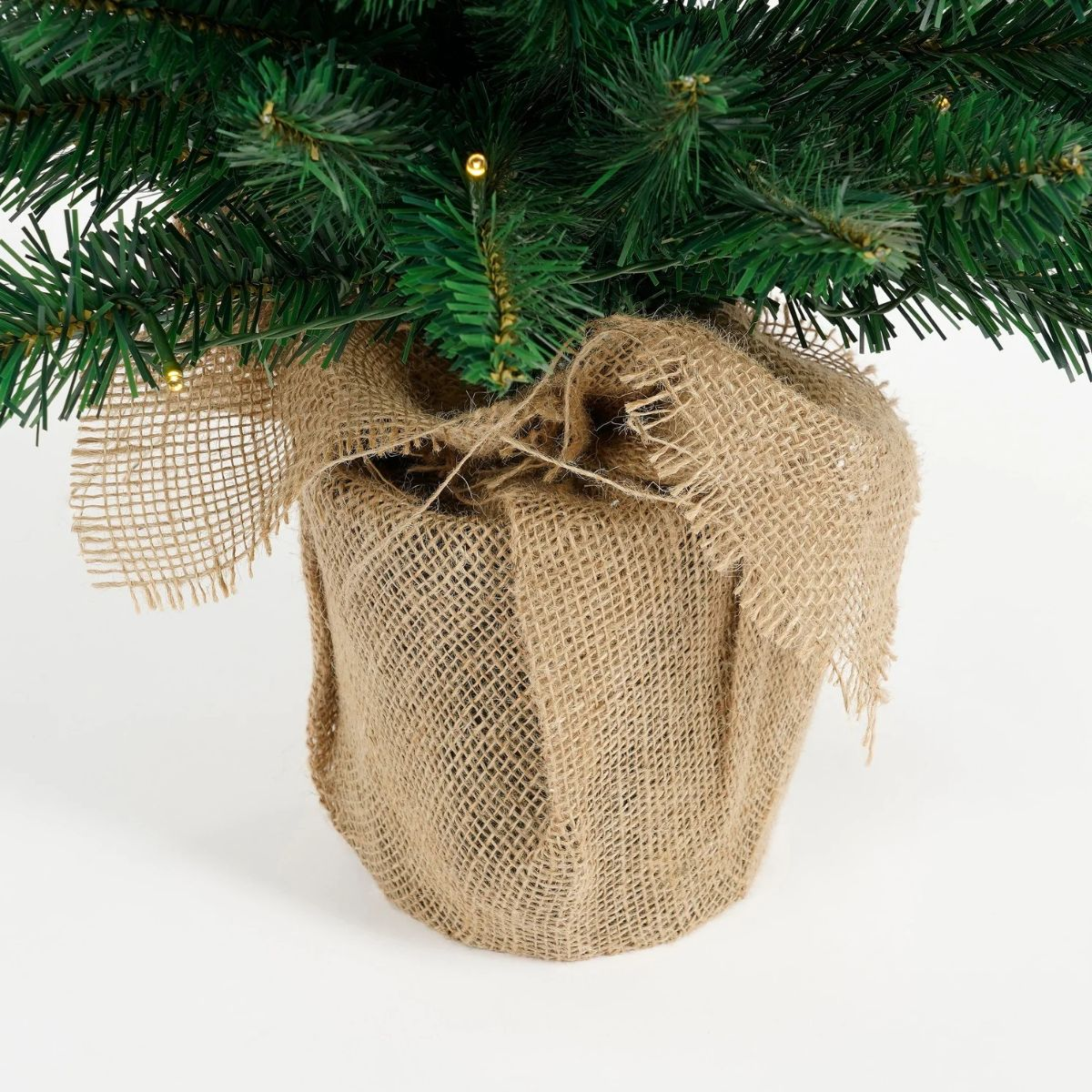 Ein kleiner künstlicher Weihnachtsbaum in einem Topf, der mit Jute umwickelt ist, vor weißem Hintergrund.