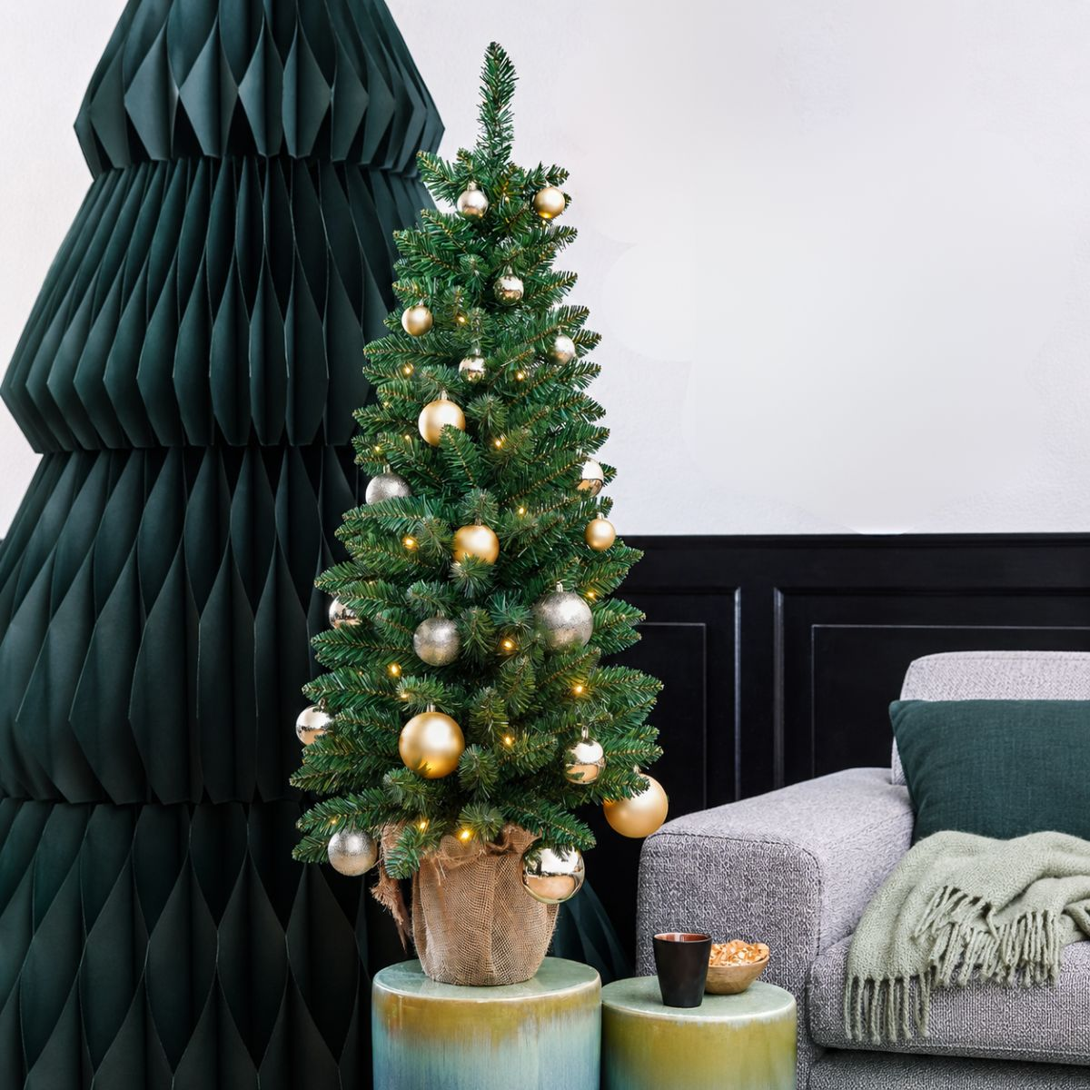 Ein kleiner Weihnachtsbaum mit goldenen und silbernen Kugeln steht auf einem Tisch neben einem grauen Sofa.