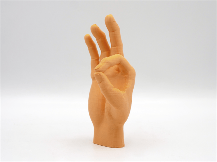 Una escultura de mano impresa en 3D de color naranja haciendo un gesto de ok sobre fondo blanco.