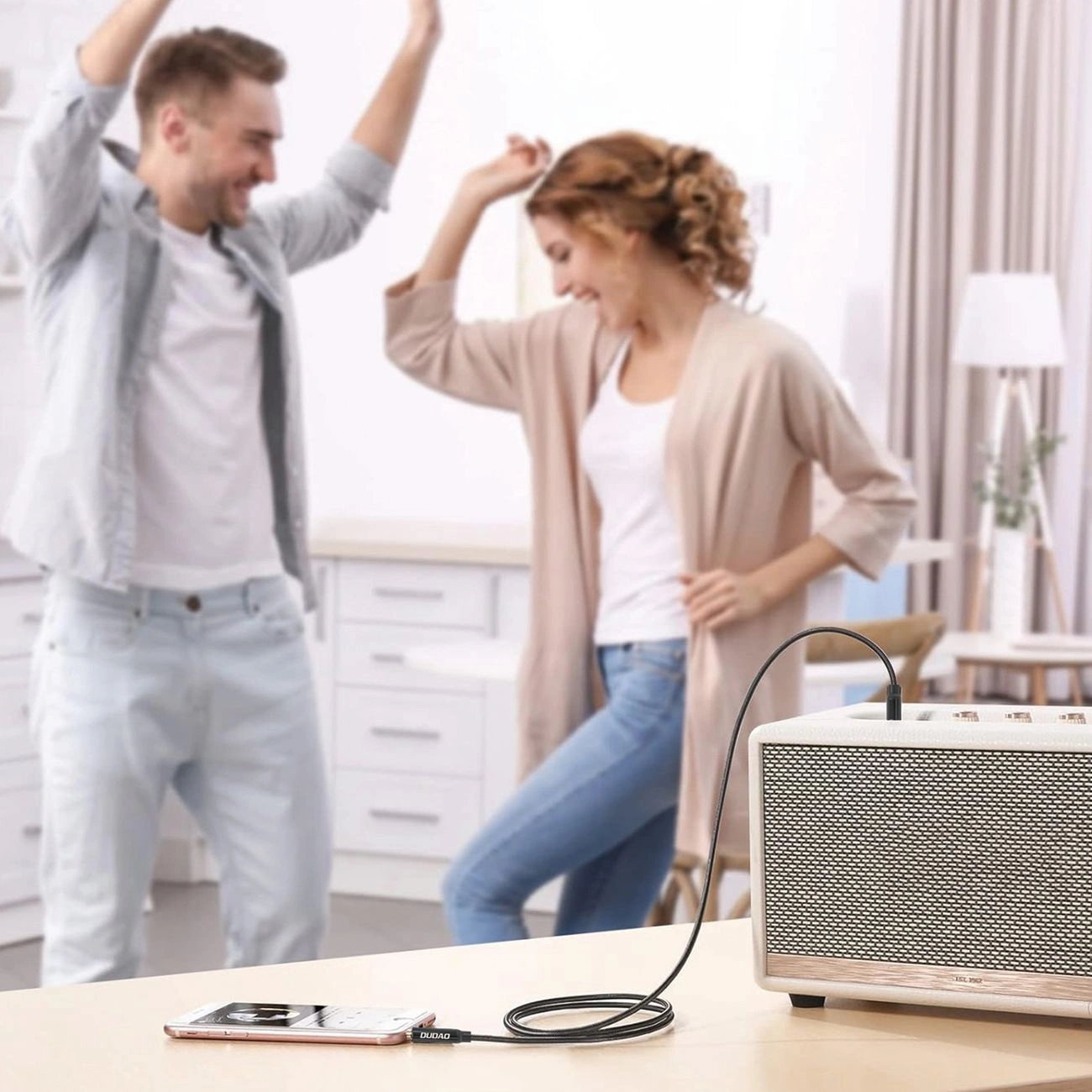 Un telefono collegato tramite cavo audio a un altoparlante bianco su un tavolo, con una coppia che balla sullo sfondo.