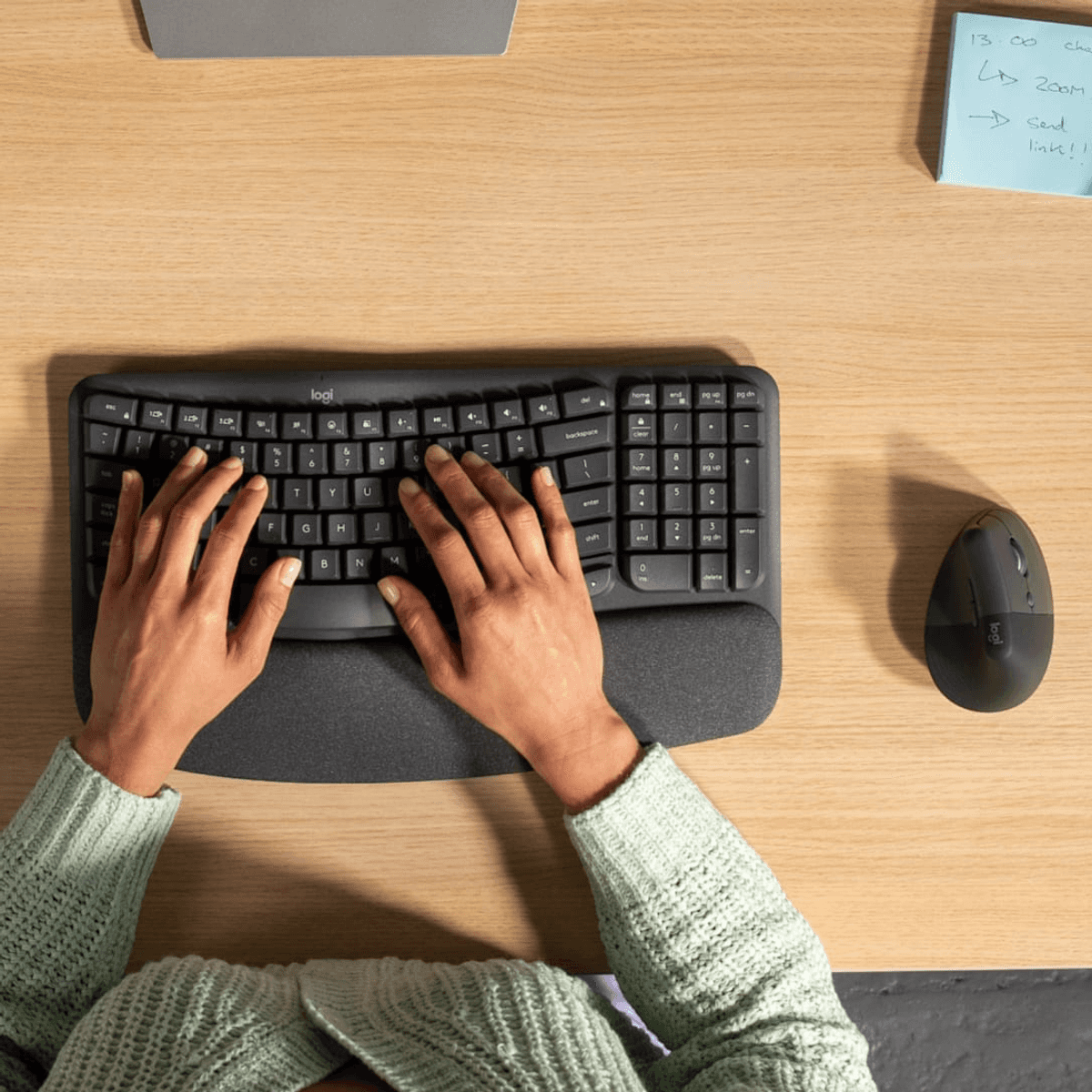 Persona che digita su una tastiera ergonomica Logitech su una scrivania in legno con mouse e nota.