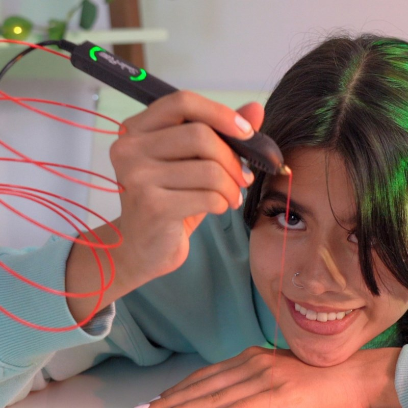 Una joven sonriente dibuja en el aire con un bolígrafo 3D rojo, sosteniendo una varita en su mano.
