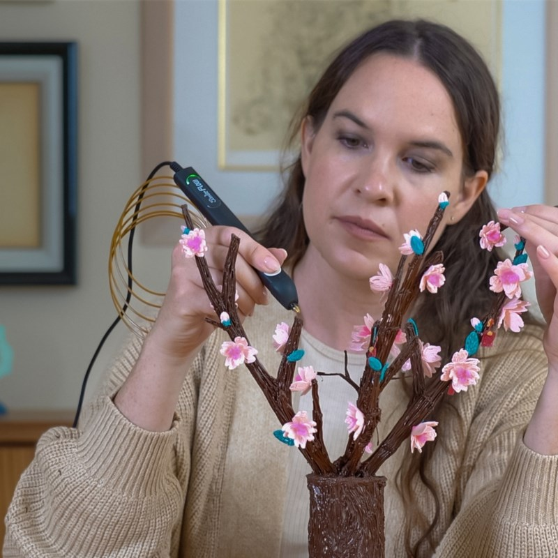 Una mujer usa un lápiz de impresión 3D para crear flores rosas en ramas.