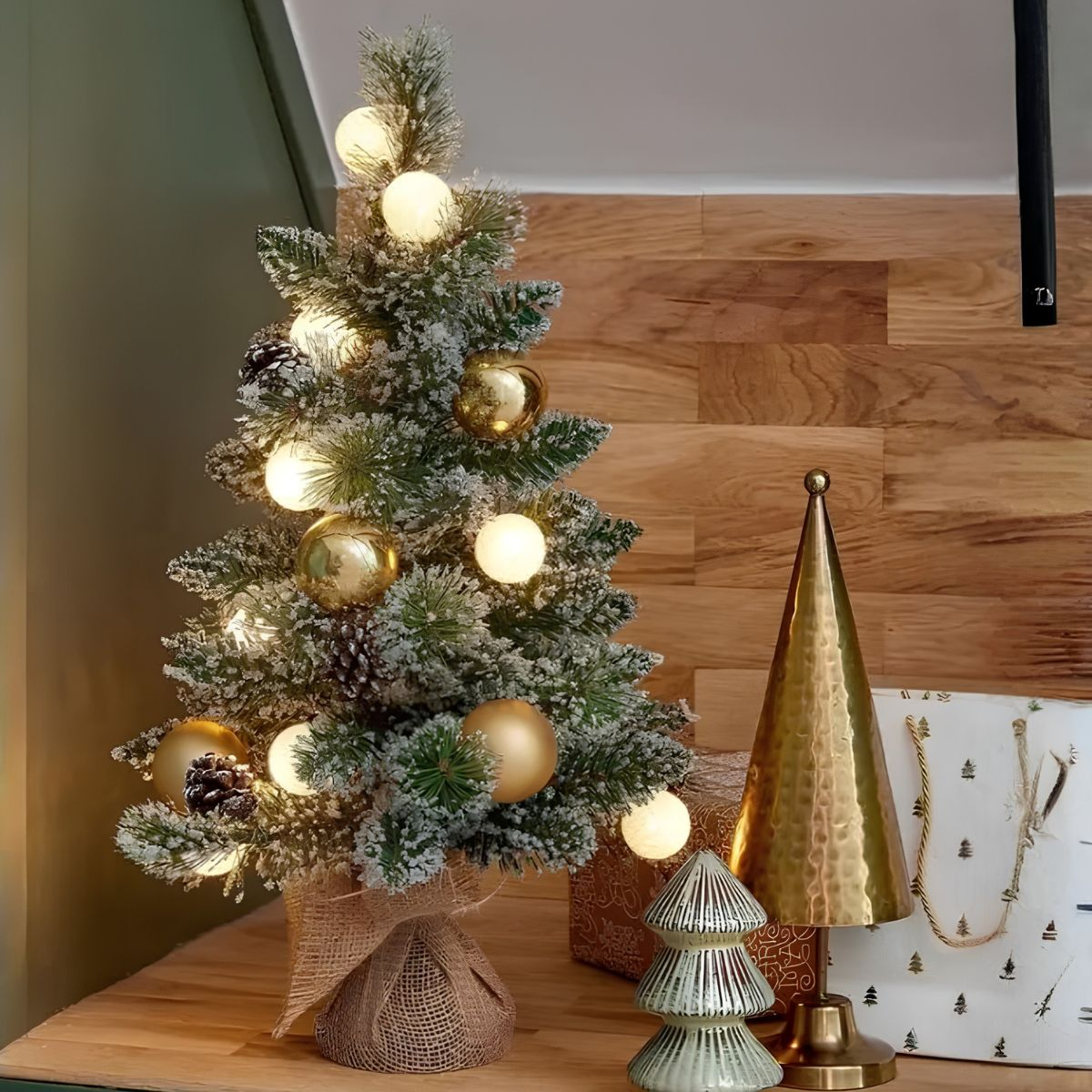 Ein geschmückter, verschneiter Tisch-Weihnachtsbaum mit Lichtern und goldenen Kugeln neben einer goldenen Tannenfigur.