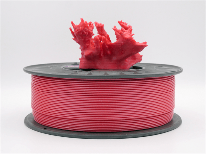 Una bobina di filamento rosso per stampante 3D con sopra una statuetta di corallo stampata rossa su sfondo chiaro.