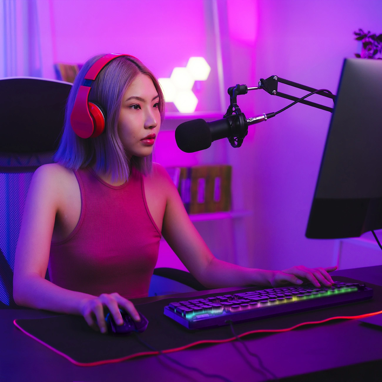 Una giovane donna con top rosso e cuffie siede al PC in una stanza illuminata di rosa.