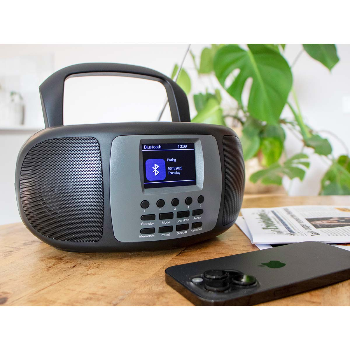 Una radio nera con schermo Bluetooth su un tavolo di legno, accanto a uno smartphone e un giornale.