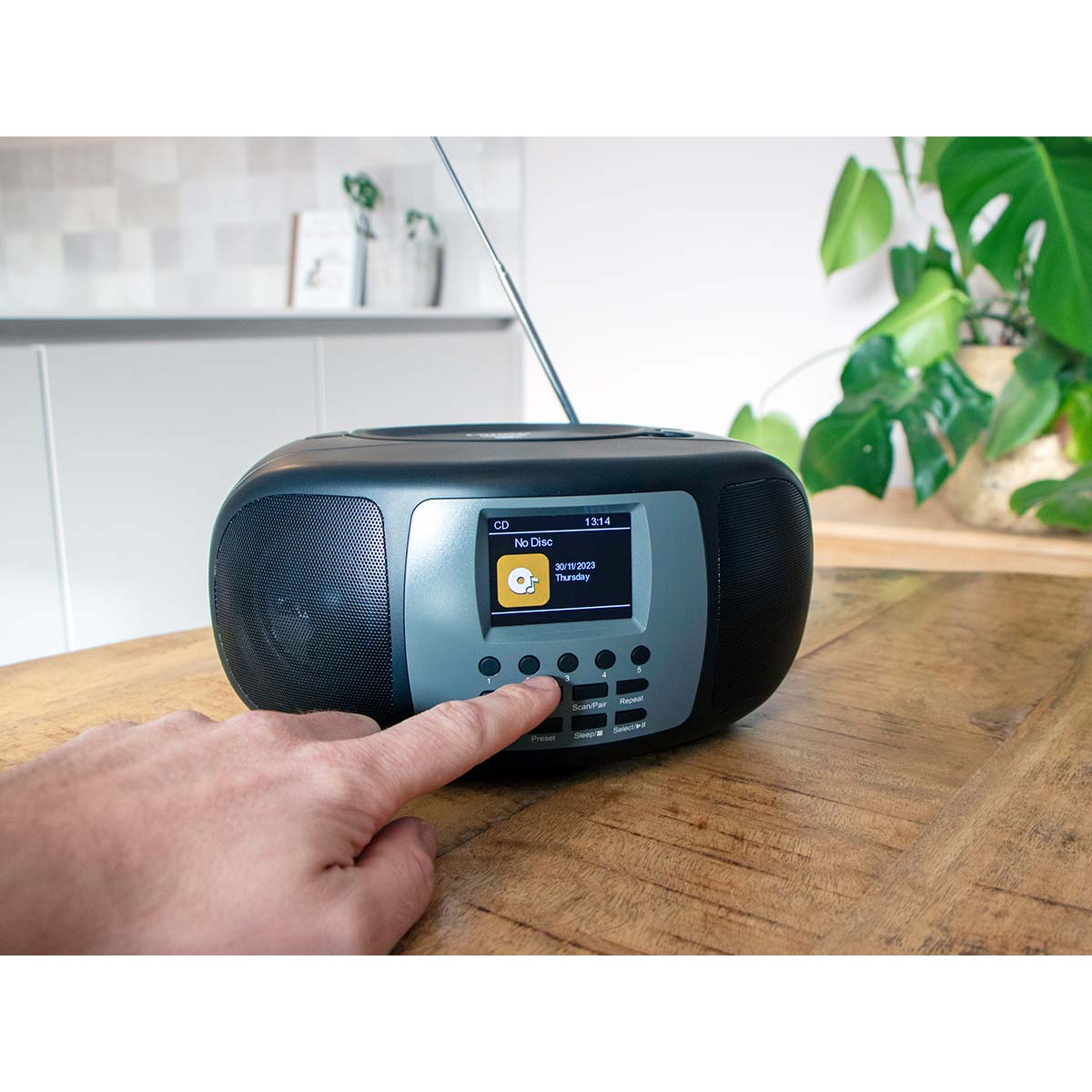 Una mano preme un pulsante su una radio grigio scuro su un tavolo di legno, con piante verdi sullo sfondo sfocato.