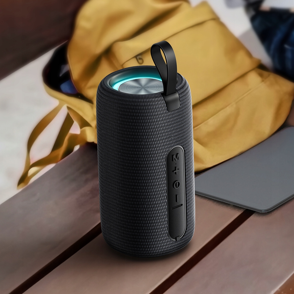 Un altoparlante Bluetooth nero cilindrico con cinturino è su una panca di legno davanti a uno zaino giallo.