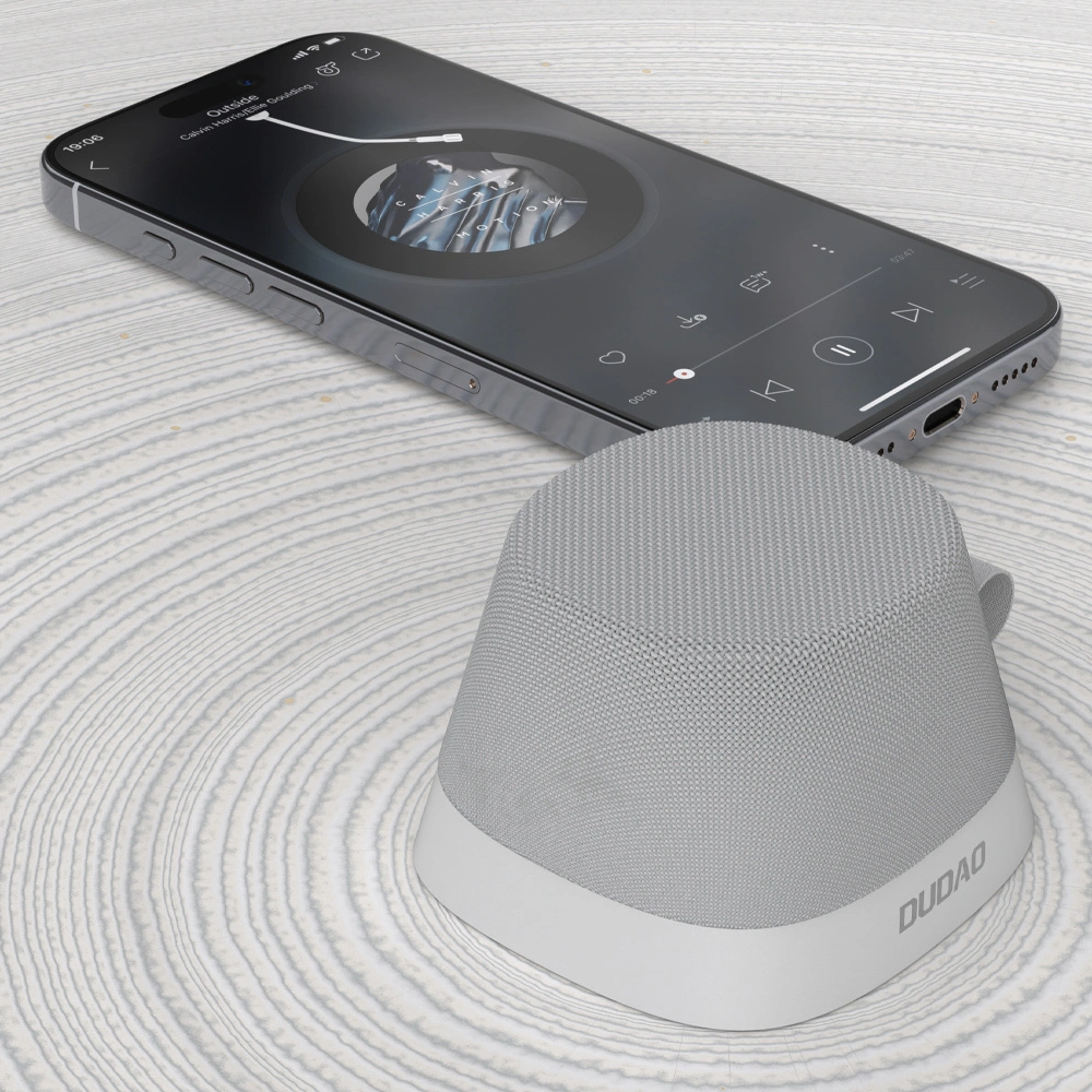 Un altoparlante wireless Dudao grigio si trova su una superficie bianca strutturata, con uno smartphone che riproduce musica sullo sfondo.