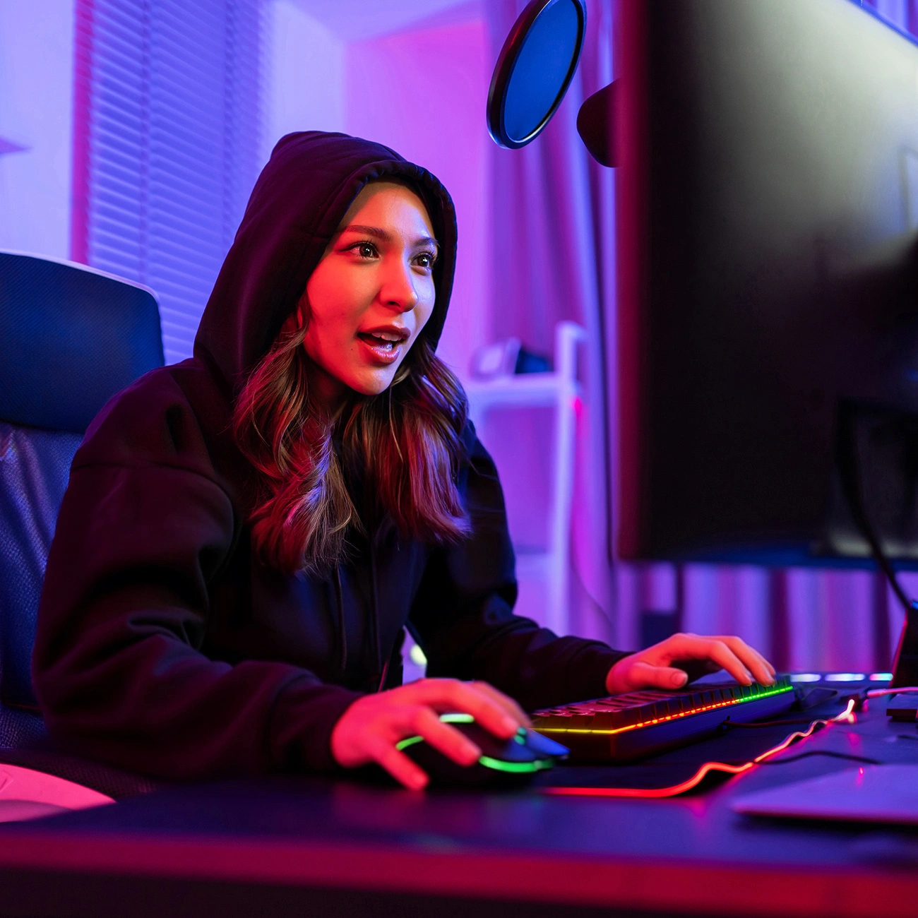 Eine junge Frau im schwarzen Hoodie sitzt in einem Raum mit neonroter und blauer Beleuchtung vor einem Computer.