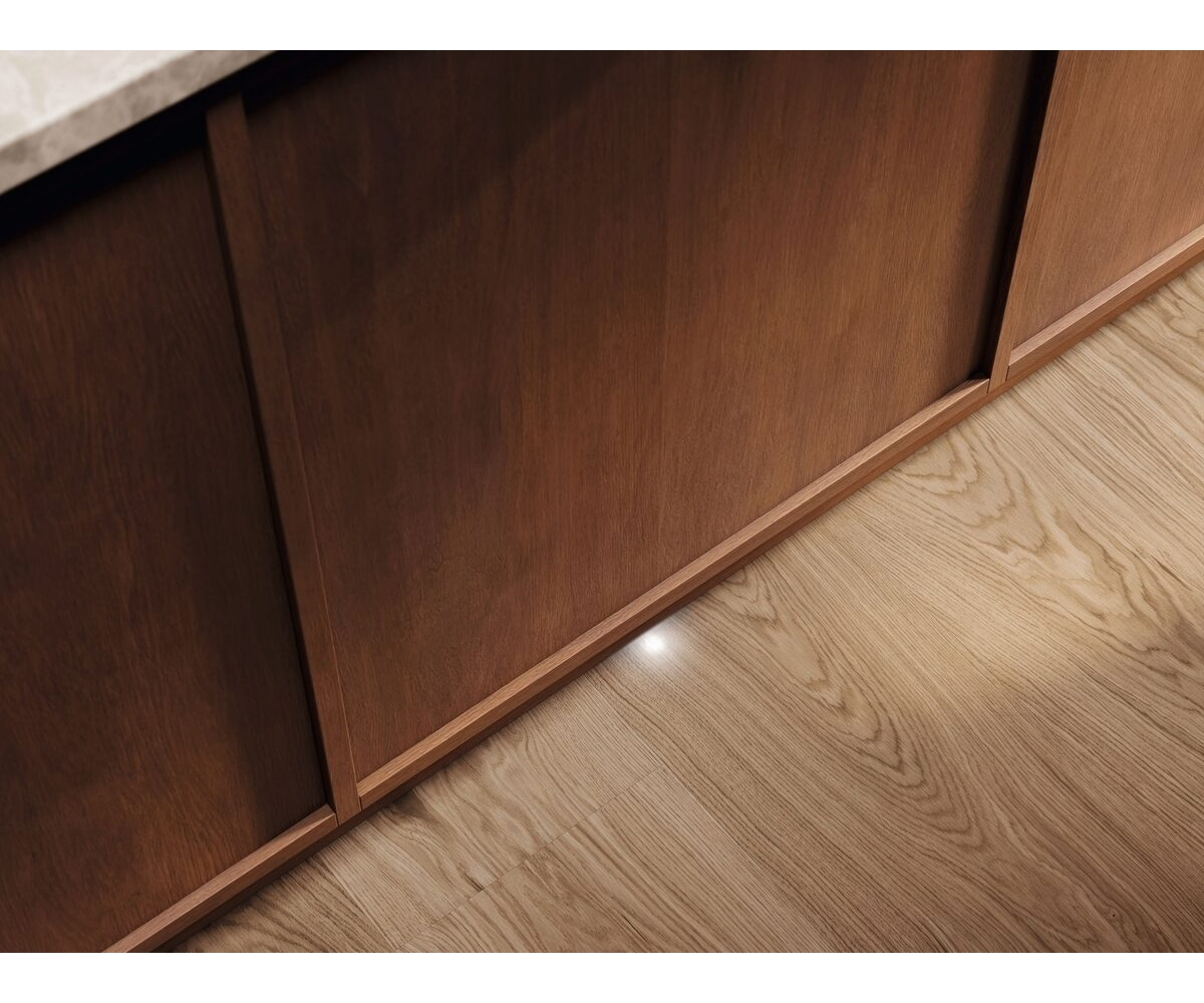 Un mobile da cucina in legno caldo con ante scorrevoli su pavimento chiaro, illuminato da una piccola luce a terra.