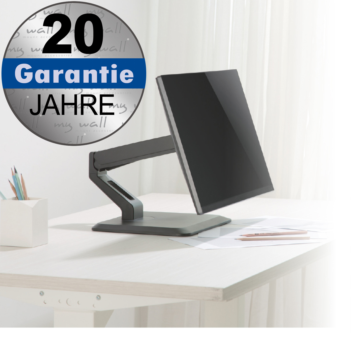 Un braccio per monitor grigio con schermo su una scrivania in legno chiaro, con un portapenne bianco a sinistra.