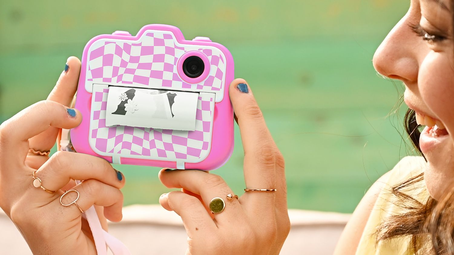 Una persona sonriente sostiene una cámara instantánea a cuadros rosa y blanco imprimiendo una foto con fondo verde.