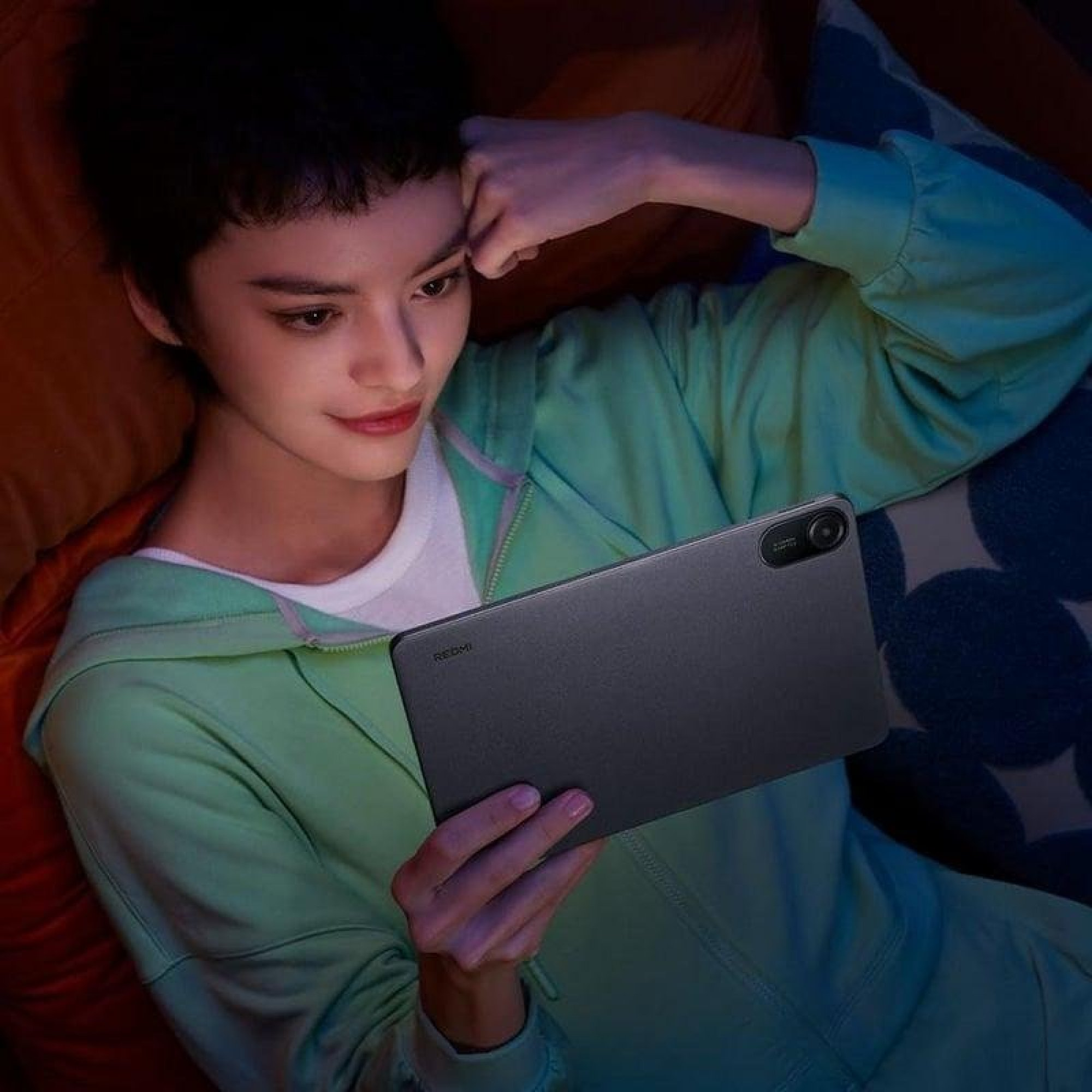 Osoba w zielonej bluzie leży na poduszkach i patrzy na szary tablet.