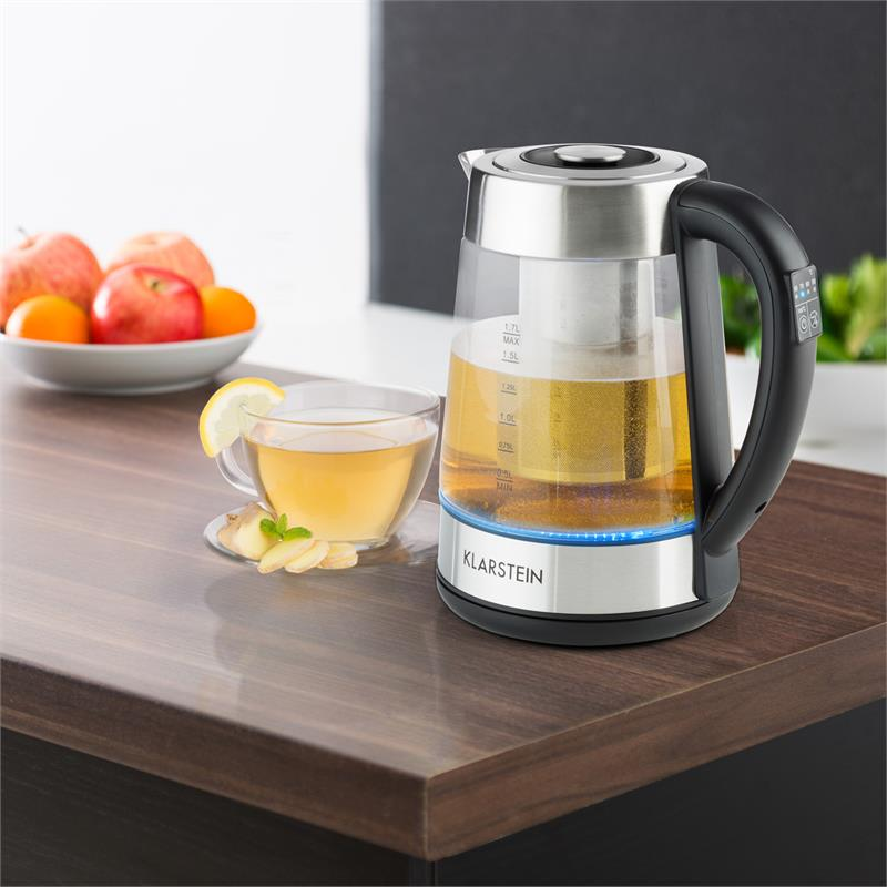 Un hervidor de cristal en un mostrador de madera con una taza de té, limón, jengibre y fruta al fondo.