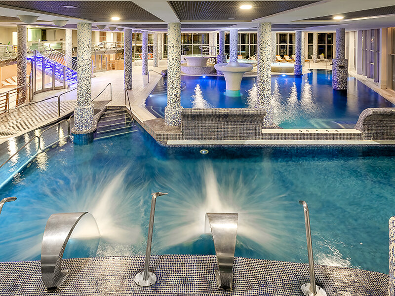 Modernas piscinas de bienestar con mosaicos, agua azul y chorros de metal en una sala luminosa con columnas.