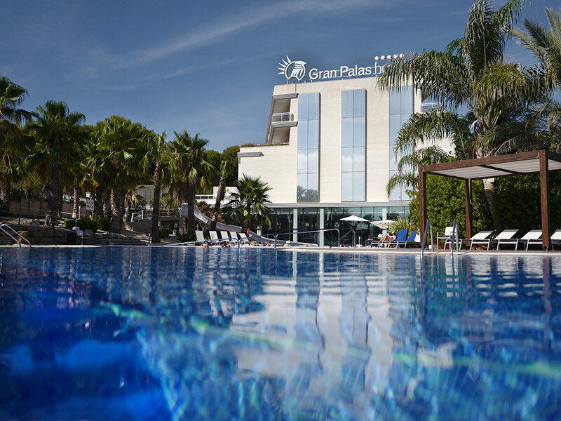 Vista exterior de un hotel de lujo con gran piscina en primer plano, palmeras y cielo azul.